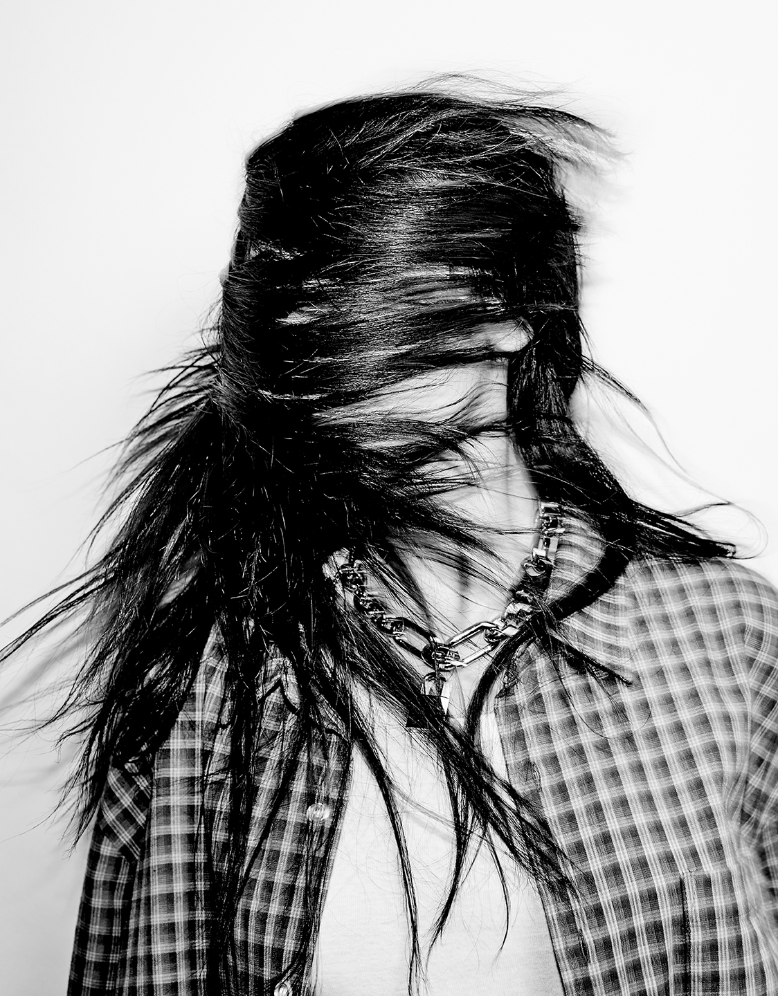 Black and white portrait of a person with long black hair blowing in their face, wearing a plaid shirt and silver chain necklace.
