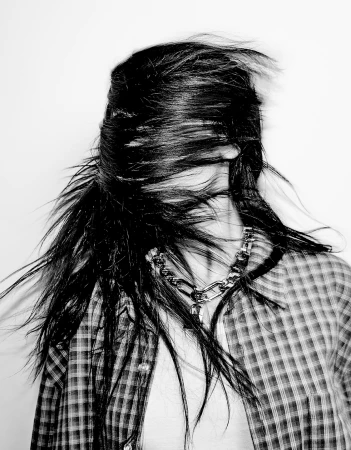 Black and white portrait of a person with long black hair blowing in their face, wearing a plaid shirt and silver chain necklace.