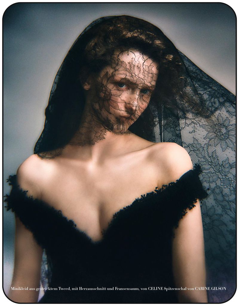Portrait of a woman wearing a black dress and a black lace veil, posing in front of a grey background in a fashion editorial.