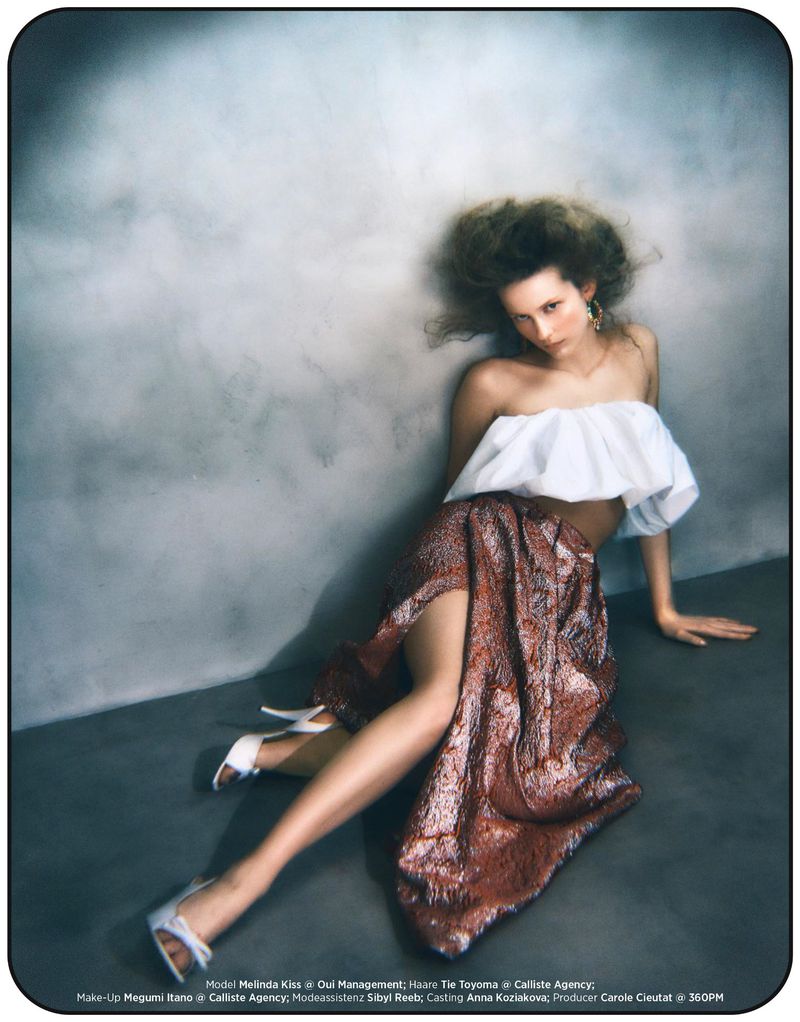Model Melinda Kiss in a white top and bronze textured skirt seated against a mottled grey studio backdrop.