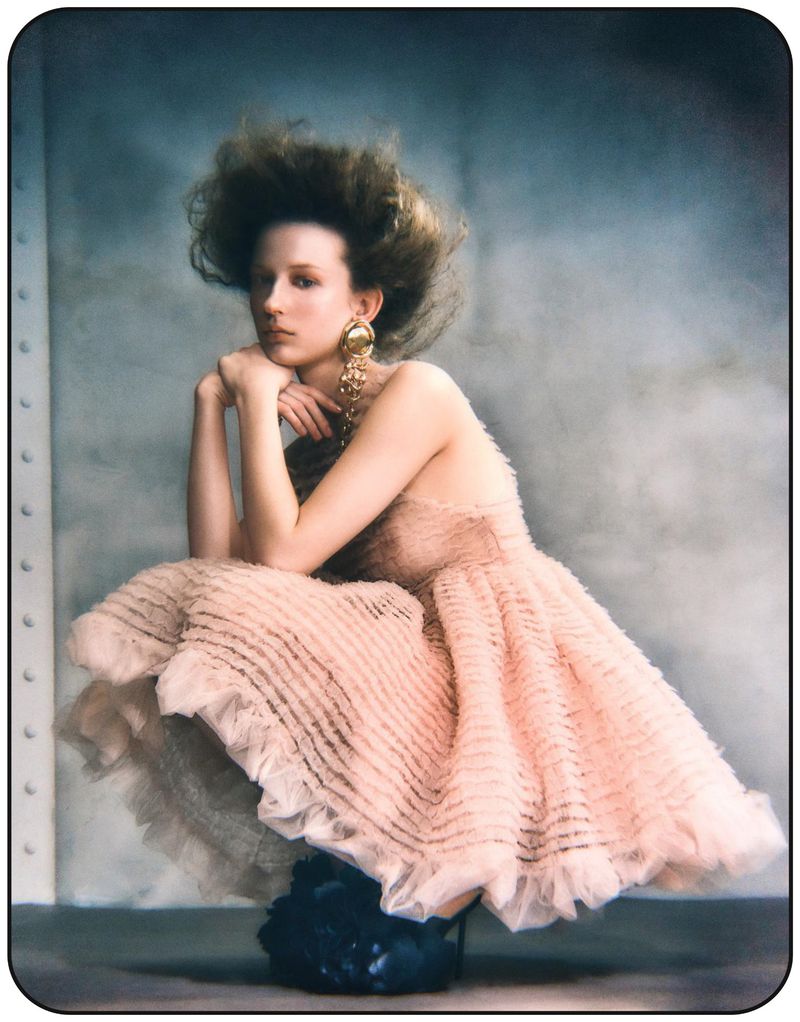 Portrait of a young woman in a strapless peach dress, crouching and resting her chin on her hands with a neutral expression, large earrings.