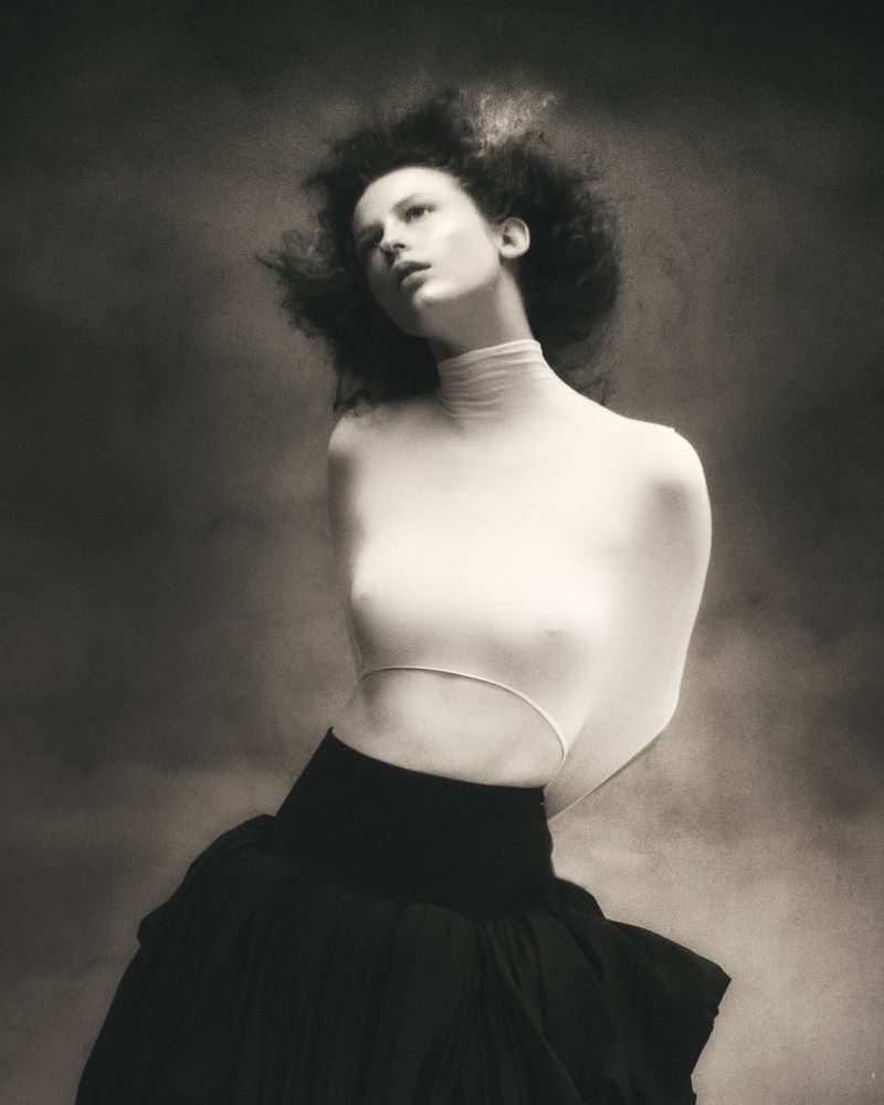 Monochrome portrait of a woman with curly hair wearing a white bodysuit and black skirt, posing in front of a cloudy background.