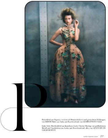 A fashion shot showcases a slender woman in an elaborate floral-embroidered dress against a soft gray background. The image is professionally styled.