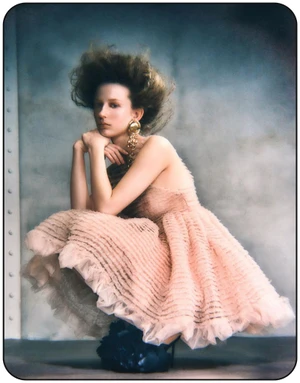 Model in ruffled pink dress with voluminous hair posing against a textured grey background for Harper's Bazaar Germany