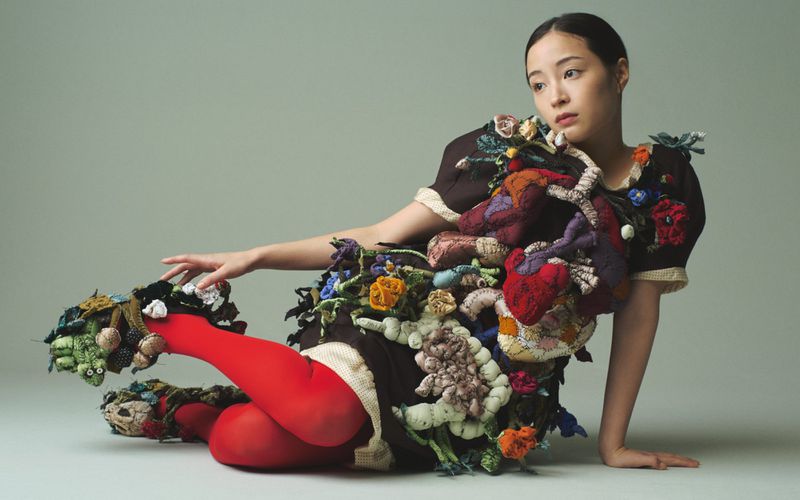 Young Asian woman in a brown dress covered in colorful crocheted flowers and red tights poses in a studio with soft, neutral lighting.