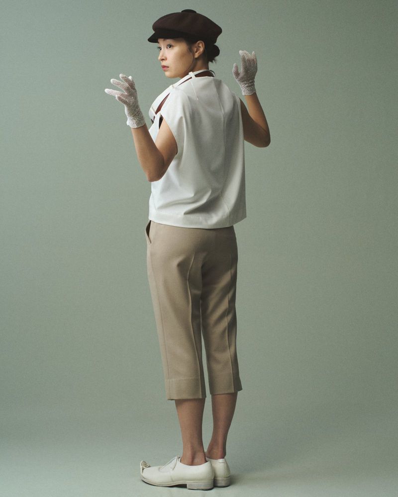 Back shot of a young Asian woman in a white top, tan cropped pants, a brown cap and white gloves, striking a curious pose in a studio.