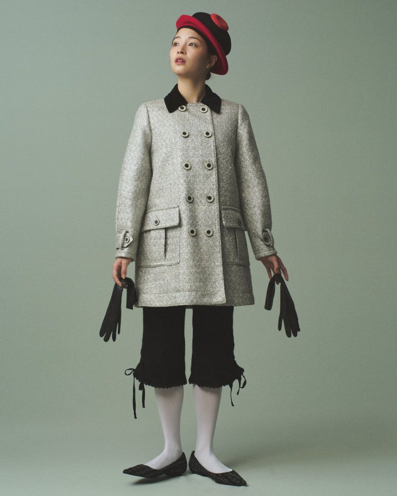 Full-length portrait of a woman in a tweed coat, black culottes with bows, white stockings, and a hat, against a plain green backdrop.