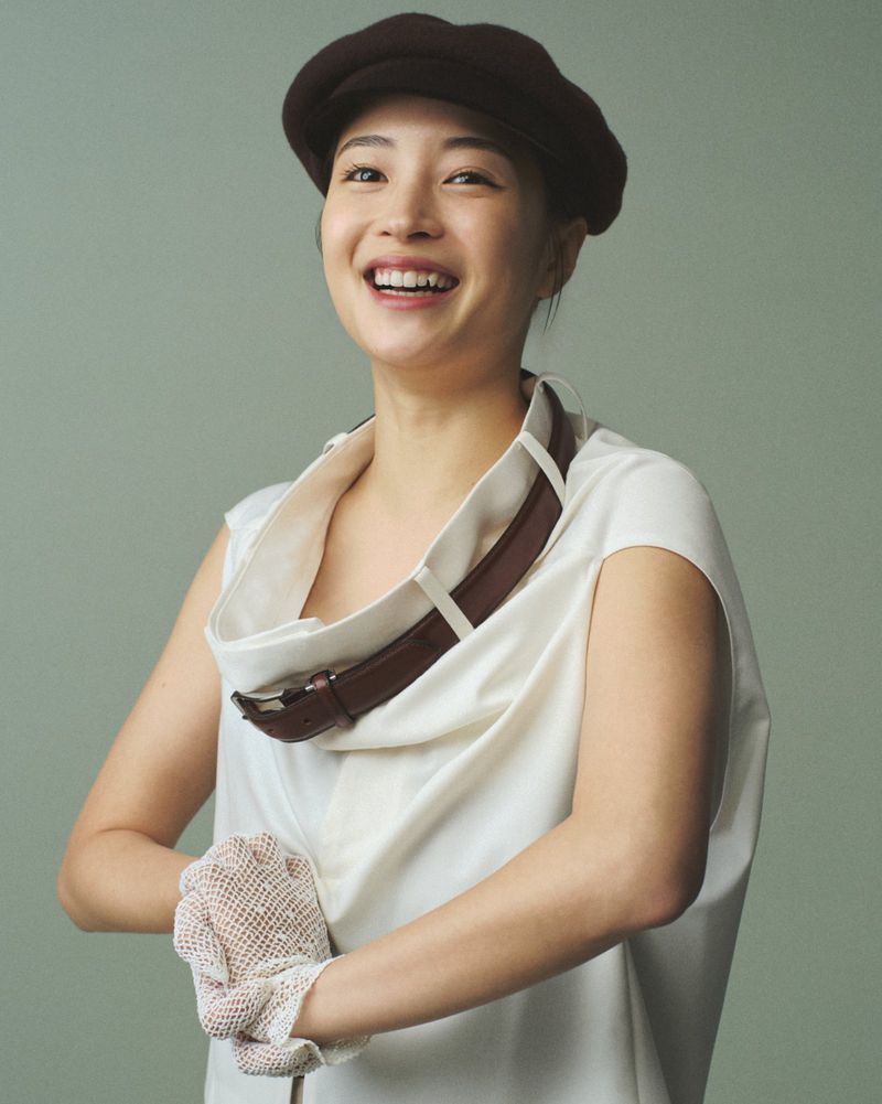 Smiling Asian woman with brown beret and ivory dress with brown belt detail, posing indoors against a solid light green background.
