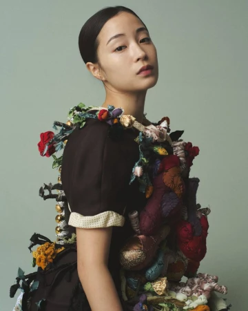 Portrait of a young adult Asian woman wearing a black dress with floral accents against a muted green background in a photo studio.