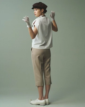 Woman wearing a white sleeveless top, tan trousers, lace gloves, and a dark cap against a grey background.