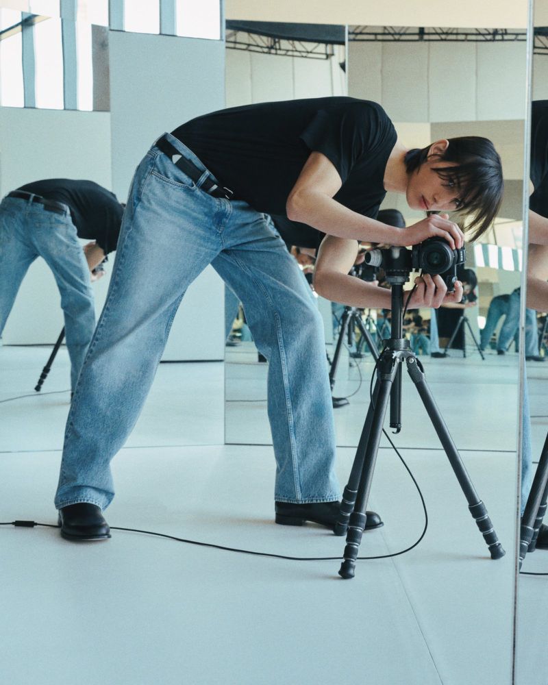 Man taking a photograph in a studio setting, using a camera mounted on a tripod, with the reflection of himself visible in mirrors.