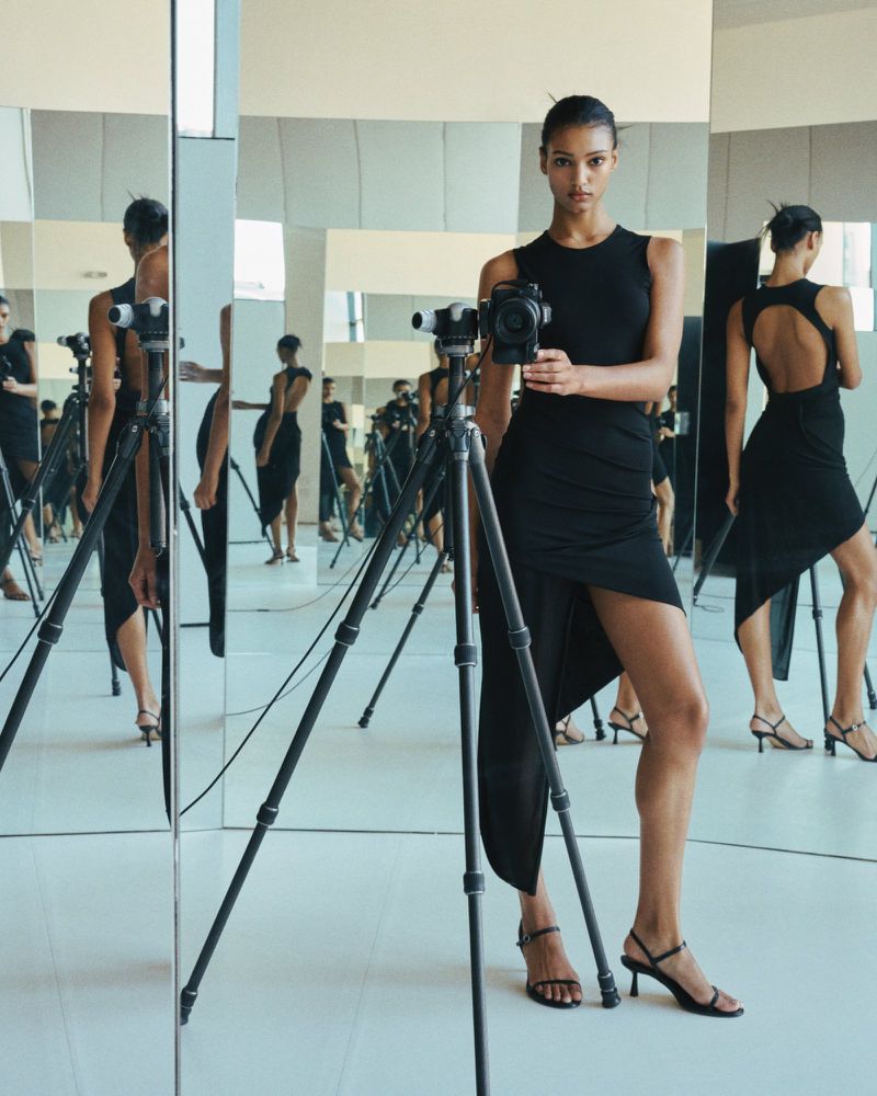 Woman in a black dress stands in front of a camera on a tripod and a mirror; studio lighting with reflective surfaces.