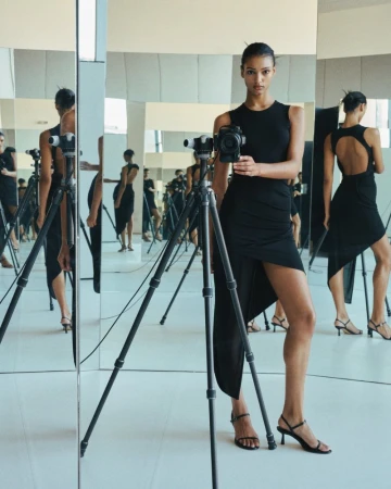 Woman in a black dress stands in front of a camera on a tripod and a mirror; studio lighting with reflective surfaces.