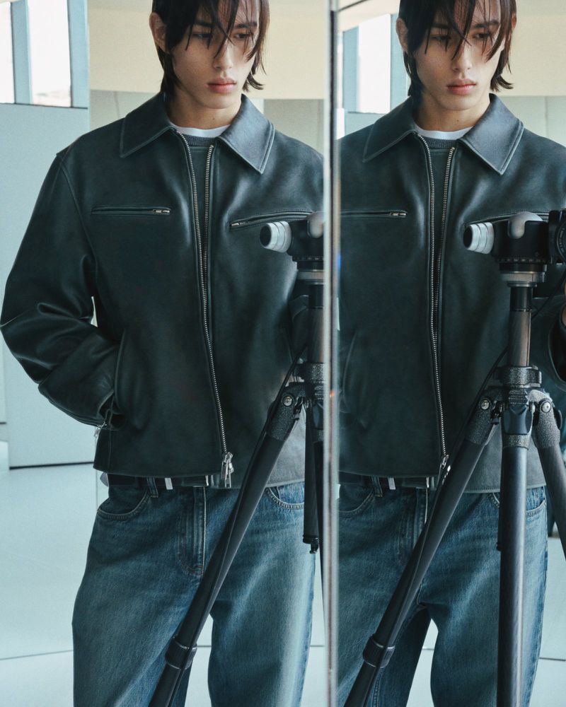 A stylish male model in a leather jacket poses in front of a mirror, holding a camera on a tripod in a bright, modern indoor setting.