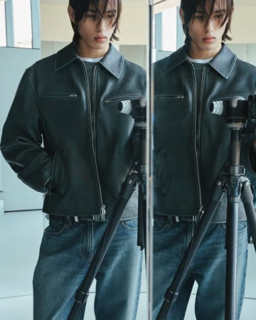 A stylish male model in a leather jacket poses in front of a mirror, holding a camera on a tripod in a bright, modern indoor setting.