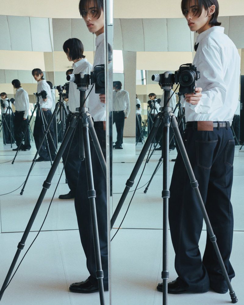 A young adult poses in front of mirrors with a camera on a tripod, wearing a white shirt and dark pants in a minimalist setting.