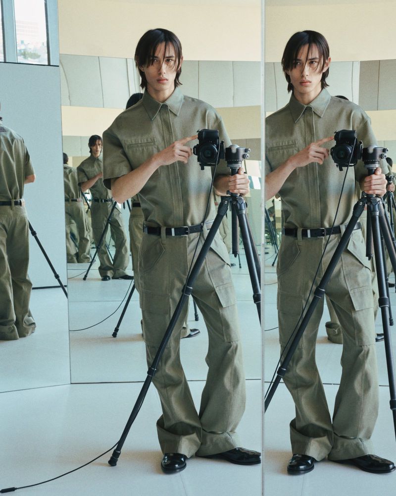 A slender man in an olive jumpsuit and black shoes poses with a vintage camera on a tripod in a bright studio setting with mirror.