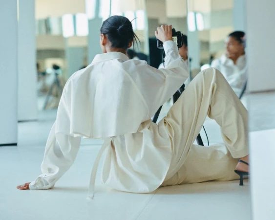 Woman in white shirt and pants poses, holding camera in a bright indoor space, reflected in a mirror, showcasing a light and airy aesthetic.