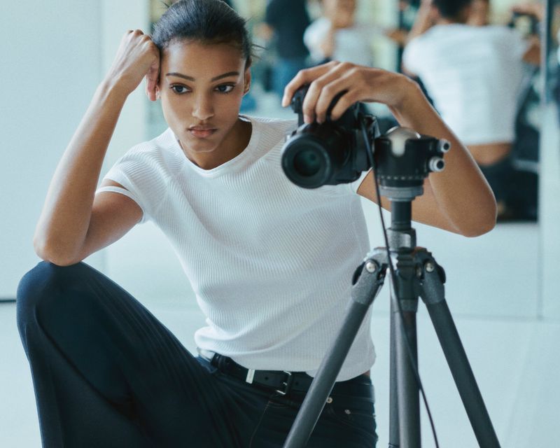 A young woman photographer with medium skin tone sits on the floor in a photo studio, reviewing images on a digital camera on a tripod.