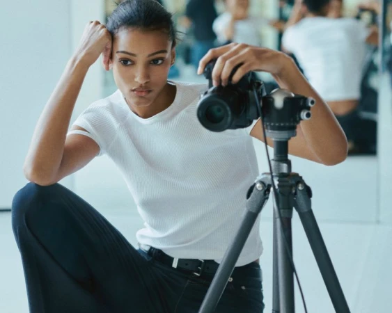 A young woman photographer with medium skin tone sits on the floor in a photo studio, reviewing images on a digital camera on a tripod.