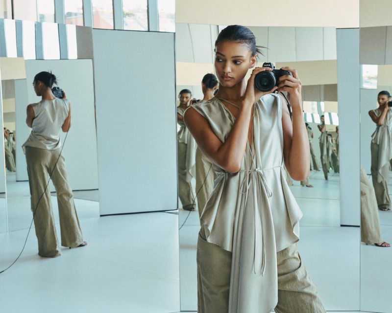 Woman in a sleeveless top and trousers takes a photo of herself in a studio with mirrors, reflecting multiple images of her.