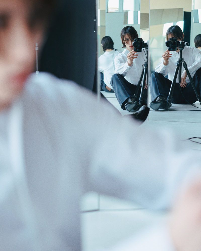 Person sits cross-legged in a reflective room, photographing themself with a DSLR camera on a tripod, creating multiple reflections.