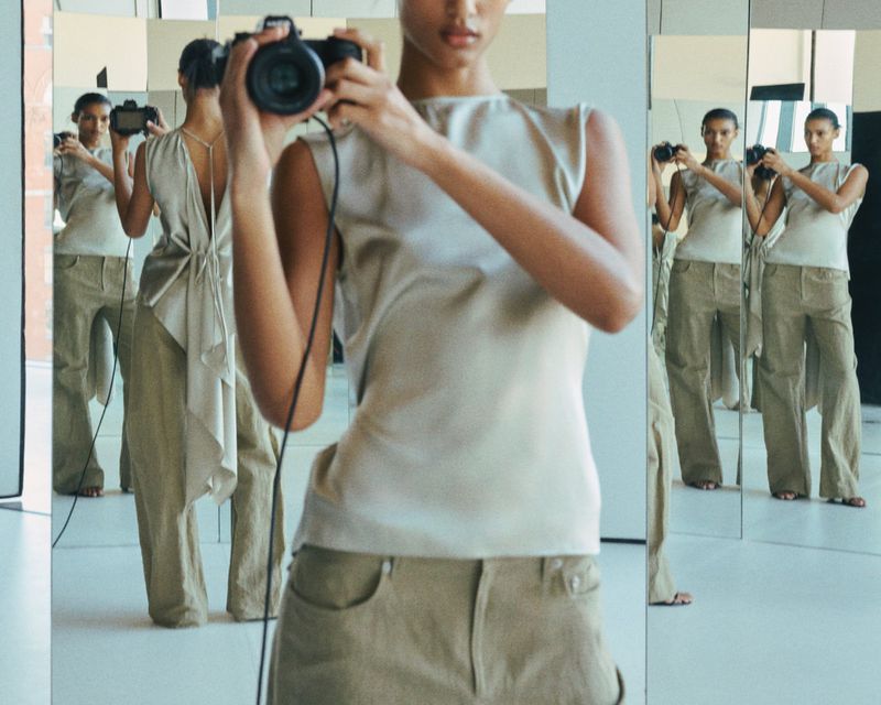 A tan-skinned woman in a beige top and pants stands in front of mirrors holding a camera up to take a photo in an indoor setting.