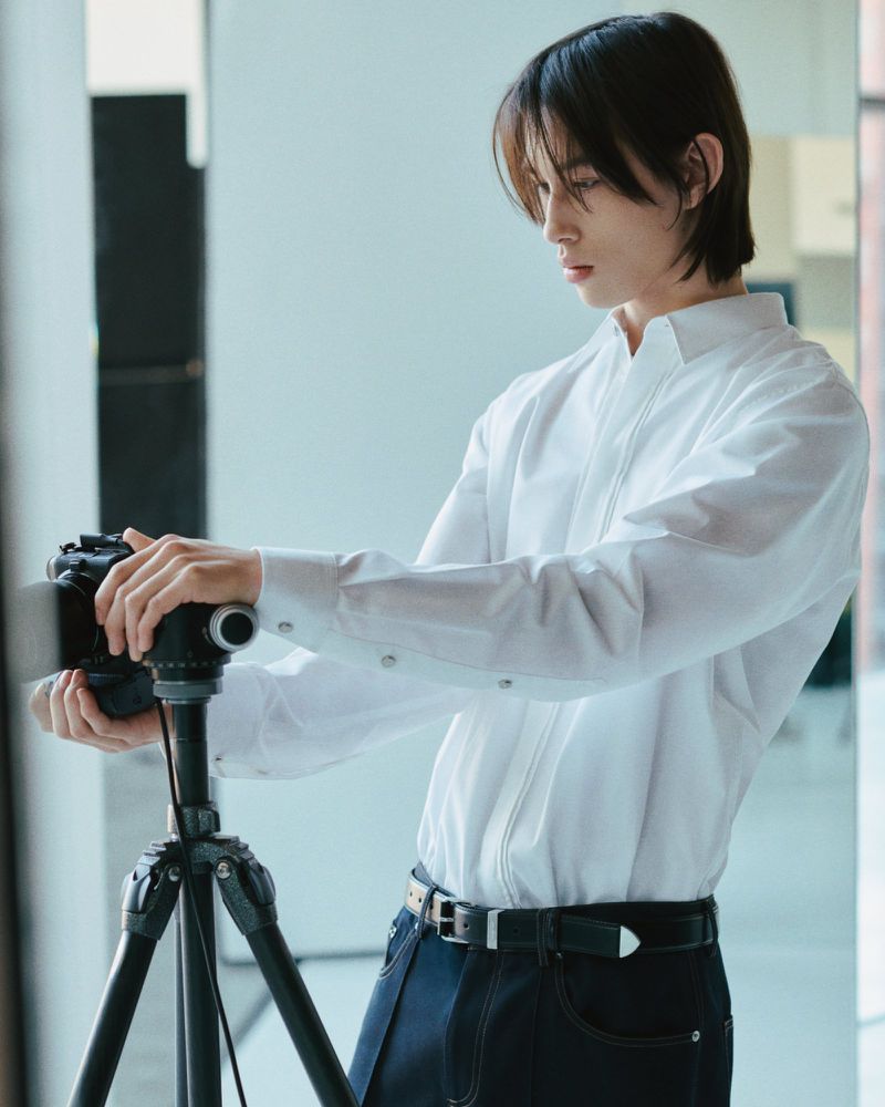 Man taking photo in studio with DSLR camera on tripod, wearing white shirt, blue jeans and black belt in a bright studio setting.