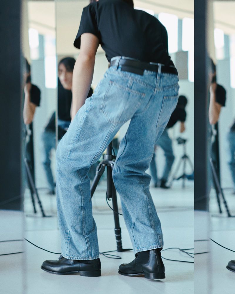 A person in jeans and black shoes stands near a camera tripod in a light-filled interior space, with reflections in mirrors.