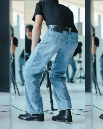 A person in jeans and black shoes stands near a camera tripod in a light-filled interior space, with reflections in mirrors.