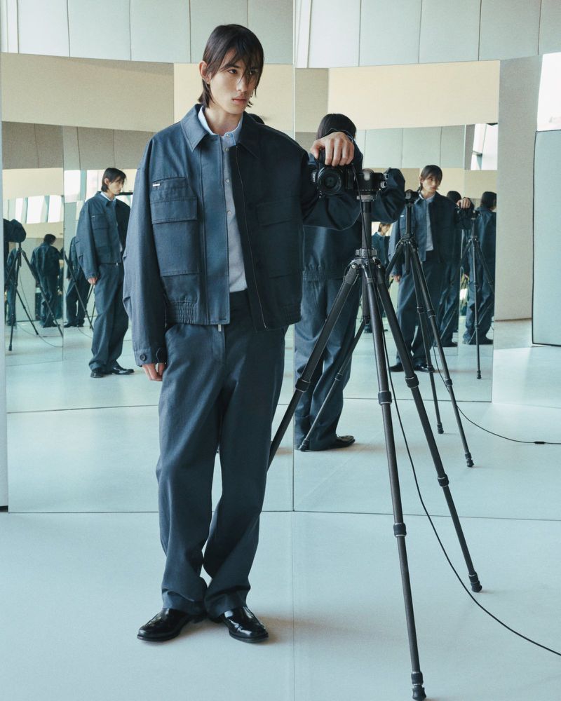 Adult male in a collared shirt, jacket, and pants poses with a camera and tripod in a bright photo studio with multiple mirrors.