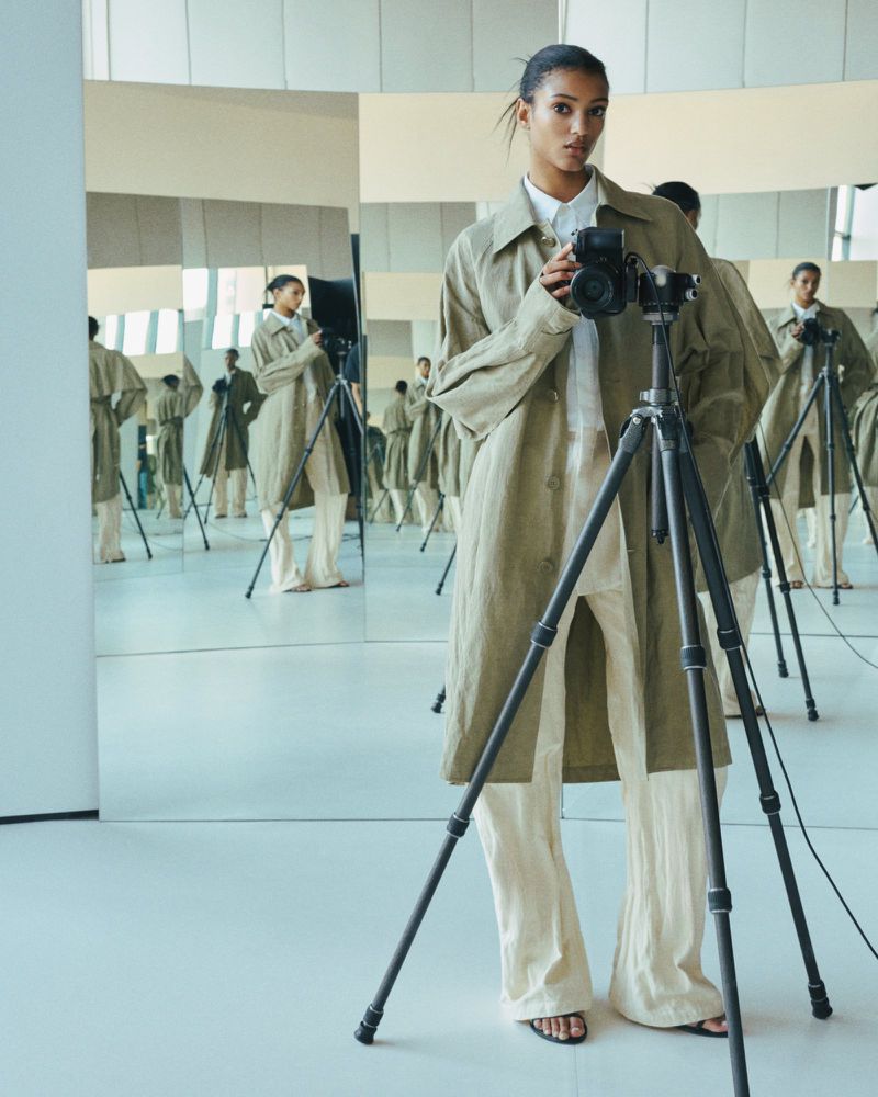 Woman holds camera on tripod in studio with mirror reflections wearing a trench coat and trousers. The scene is well-lit with neutral colors.