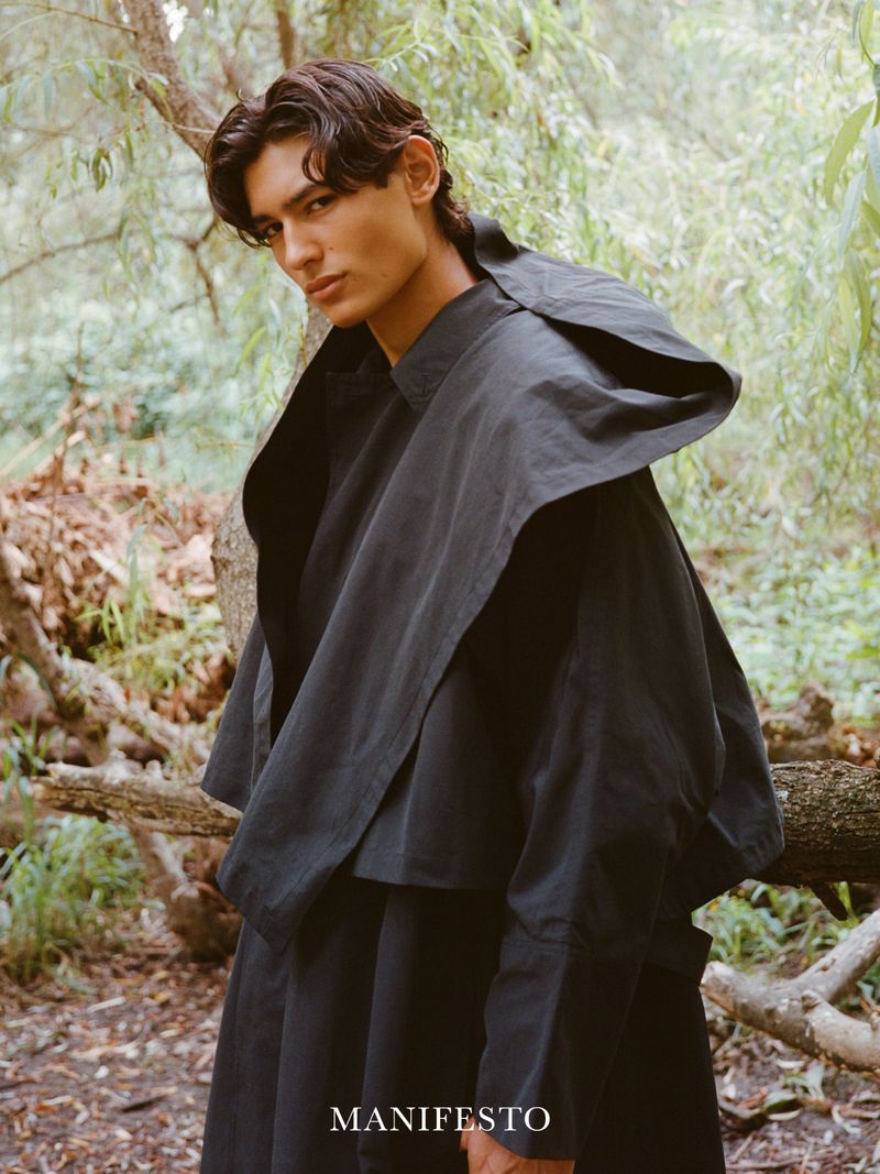 Portrait of a young adult man in a dark hooded garment, standing outdoors among trees in a fashion editorial style.
