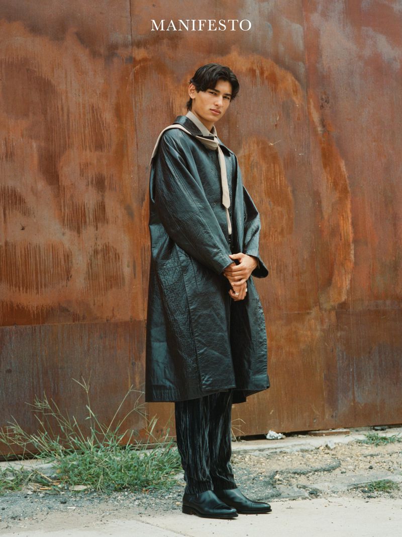 Male fashion model posing in a black coat and matching pants against a rusty metal wall with sparse ground vegetation.