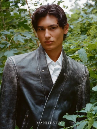 Model wearing a dark distressed leather jacket and white shirt posed amidst lush green foliage in a woodland setting.