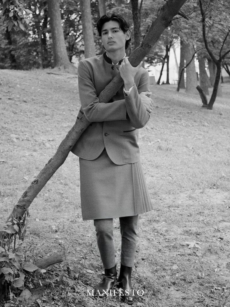 Black and white portrait of a male in a blazer, skirt, and dress pants leaning against a tree in a park-like setting.