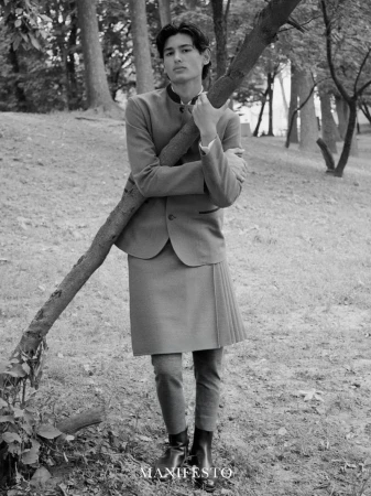 Black and white portrait of a male in a blazer, skirt, and dress pants leaning against a tree in a park-like setting.