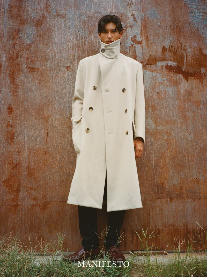 Adult male fashion model poses in long cream overcoat, dark pants, and brown dress shoes in front of a textured brown metal wall outdoors.