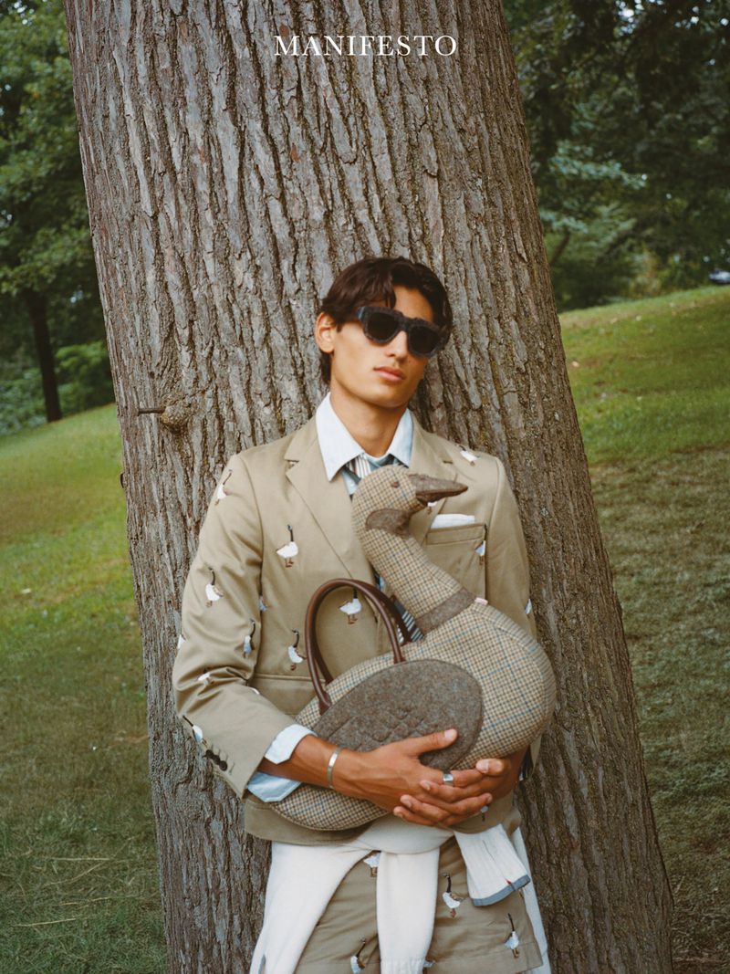 Man in a patterned tan suit holding a large duck-shaped purse stands near a tree in an outdoor setting with greenery.