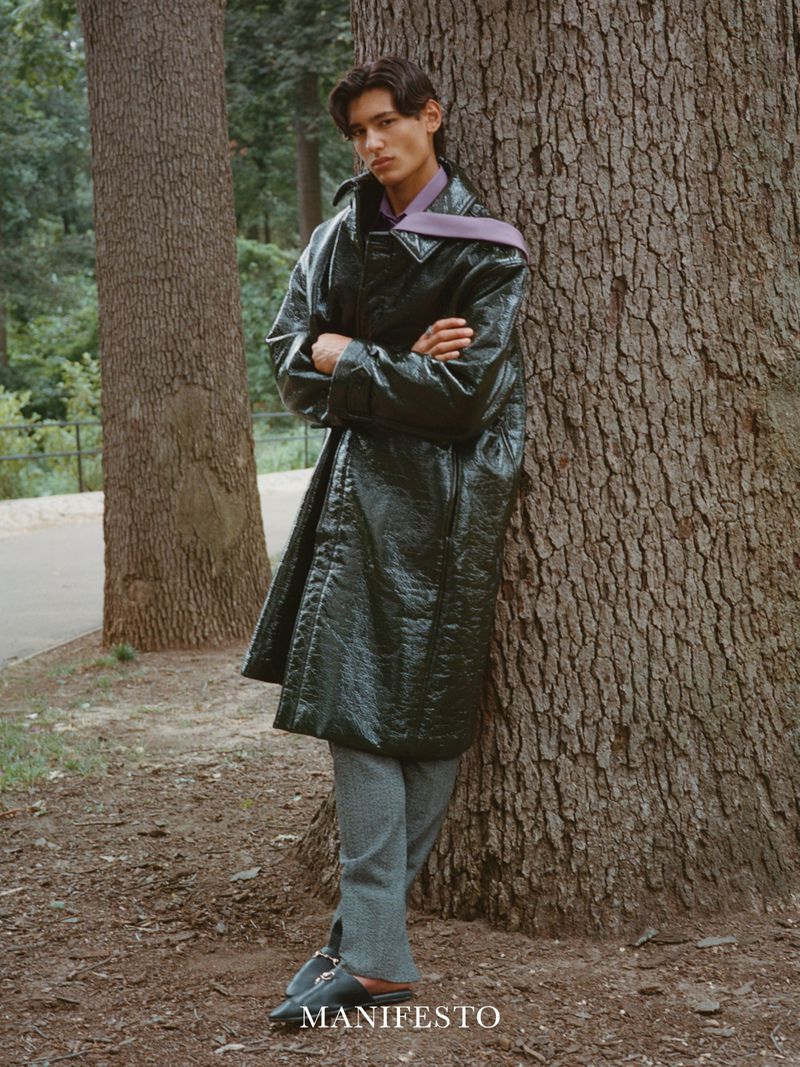Male model posing next to a tree in a forest wearing a long, green coat, gray pants, and black shoes for a fashion editorial.