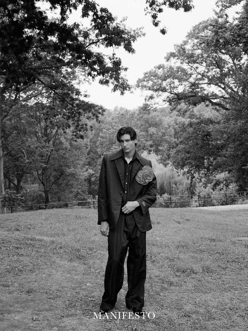 Man wearing a dark suit with a floral lapel pin standing outdoors in a wooded park in a black and white photograph.