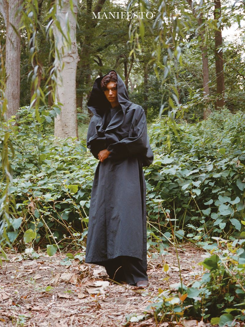 Young adult stands in lush green forest wearing flowing dark hooded cape with soft natural light filtering through the dense foliage.