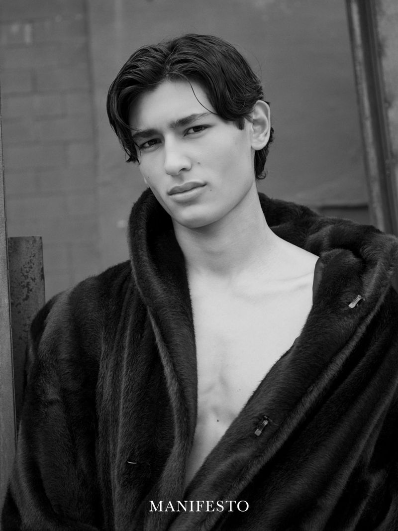 Black and white portrait of a young adult male with dark hair, wearing a fur coat, posing in an outdoor setting against a textured wall.
