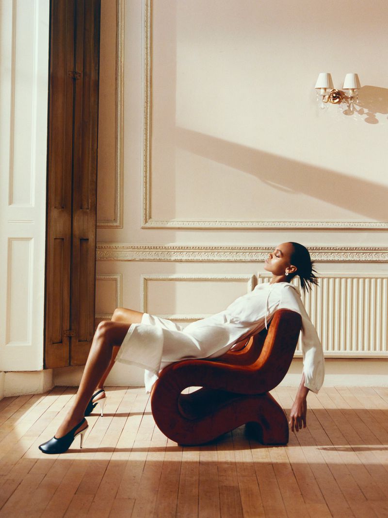 A woman lounges in a modern rust-colored chair in a sunlit room, wearing a white dress and black heels, creating an elegant fashion portrait.