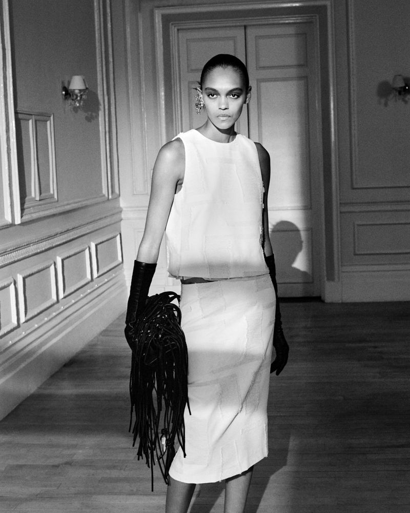 Elegant studio shot of a Black woman in a white skirt suit, accessorized with long black gloves and unique fringe purse, against a formal backdrop.