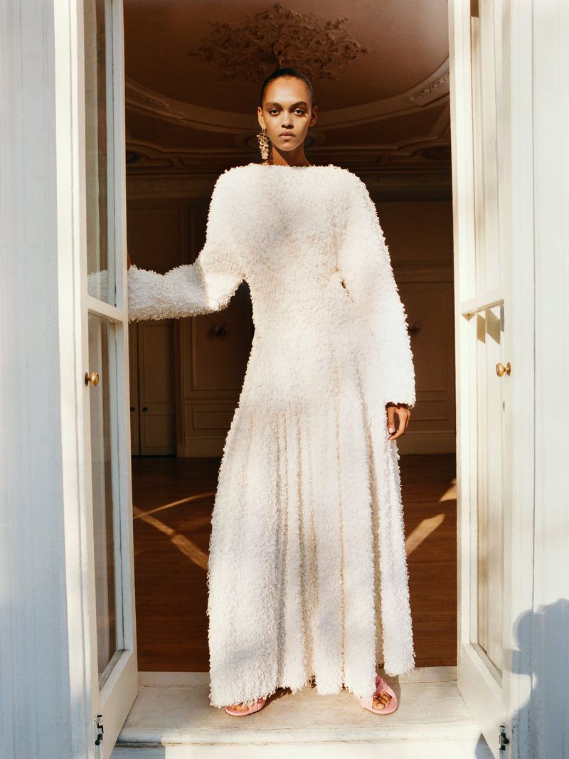 A woman poses in a full-length white dress in a doorway with natural light, showcasing a fashion editorial style.