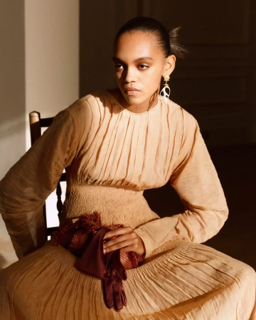 Elegant young woman in a flowing beige dress sits on a wooden chair in soft light, accented by dark red gloves and statement earrings.