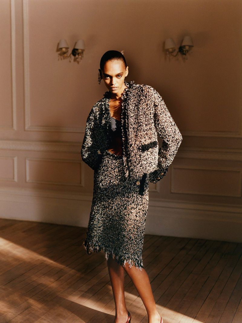 An elegant adult woman poses in a tweed suit with a jacket and knee-length skirt indoors on a wooden floor in front of a pale pink wall.