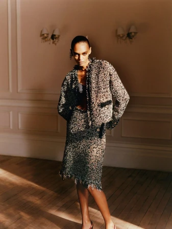 An elegant adult woman poses in a tweed suit with a jacket and knee-length skirt indoors on a wooden floor in front of a pale pink wall.
