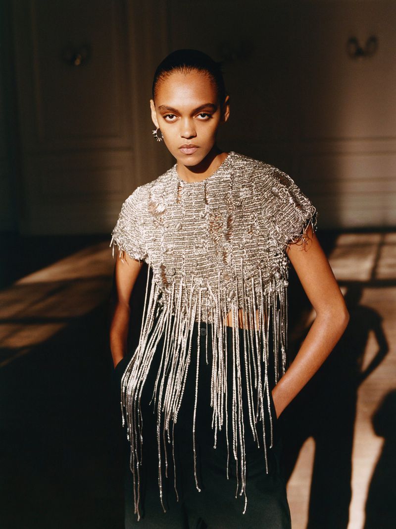 Fashion shot of a Black female model wearing a silver beaded top with fringe and black pants, posing indoors with a neutral background.